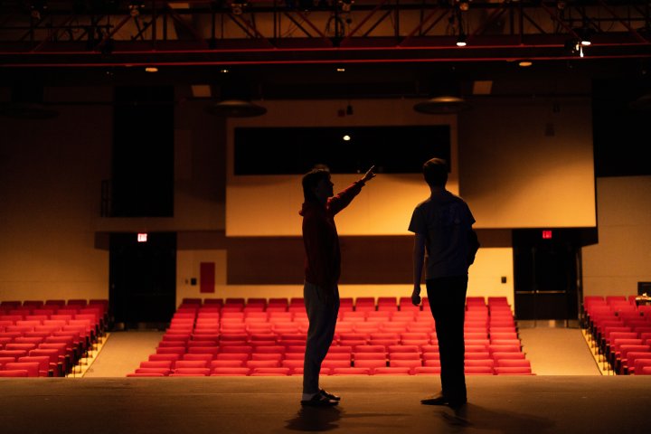 Two figures stand on a dimly lit stage, silhouetted against a backdrop of empty red theater seats. One person points toward the audience, while the other observes. The scene captures a quiet moment of rehearsal or preparation in the auditorium.
